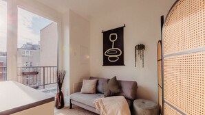 A modern living room with a sofa, a small round ottoman, a decorative wall piece, and a large window offering a view of buildings.