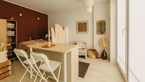 Une pièce compacte et bien éclairée, comprenant un lit, un petit bureau avec des chaises, une bibliothèque et un miroir.
