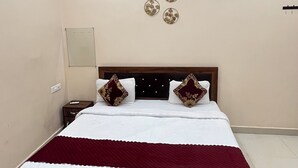A neatly made bed with a dark wooden frame, white bedding, and two decorative pillows. There is a ceiling fan above the bed, and the room has plain walls with a small mirror and three wall-mounted light fixtures. - R K Lodge And Palace  (Ayodhya)