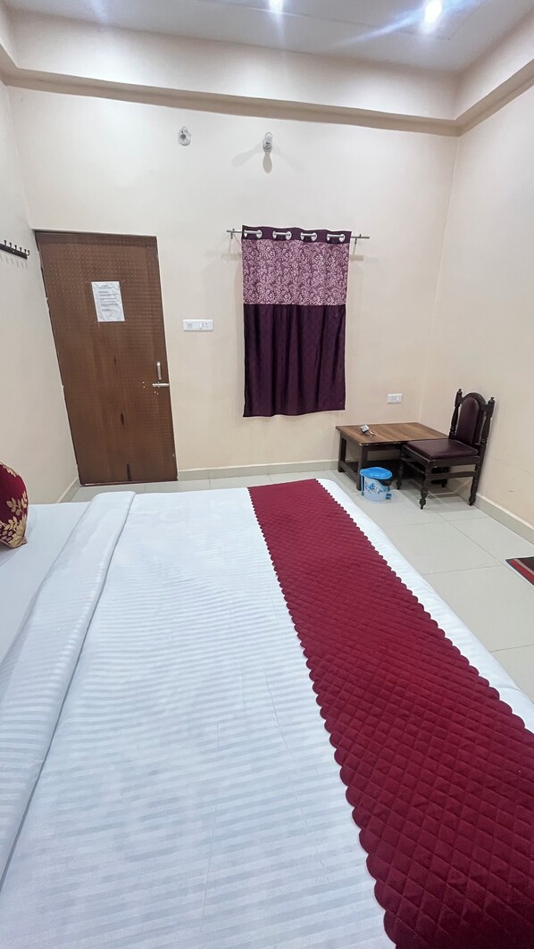 A bedroom with a bed, a wooden door, a window with a curtain, a small table, and a chair. - R K Lodge And Palace (Ayodhya)