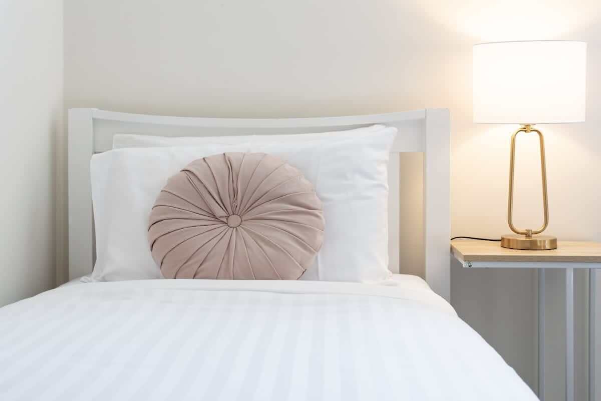 A bed with a white headboard, a beige decorative pillow, and a bedside table with a lamp.