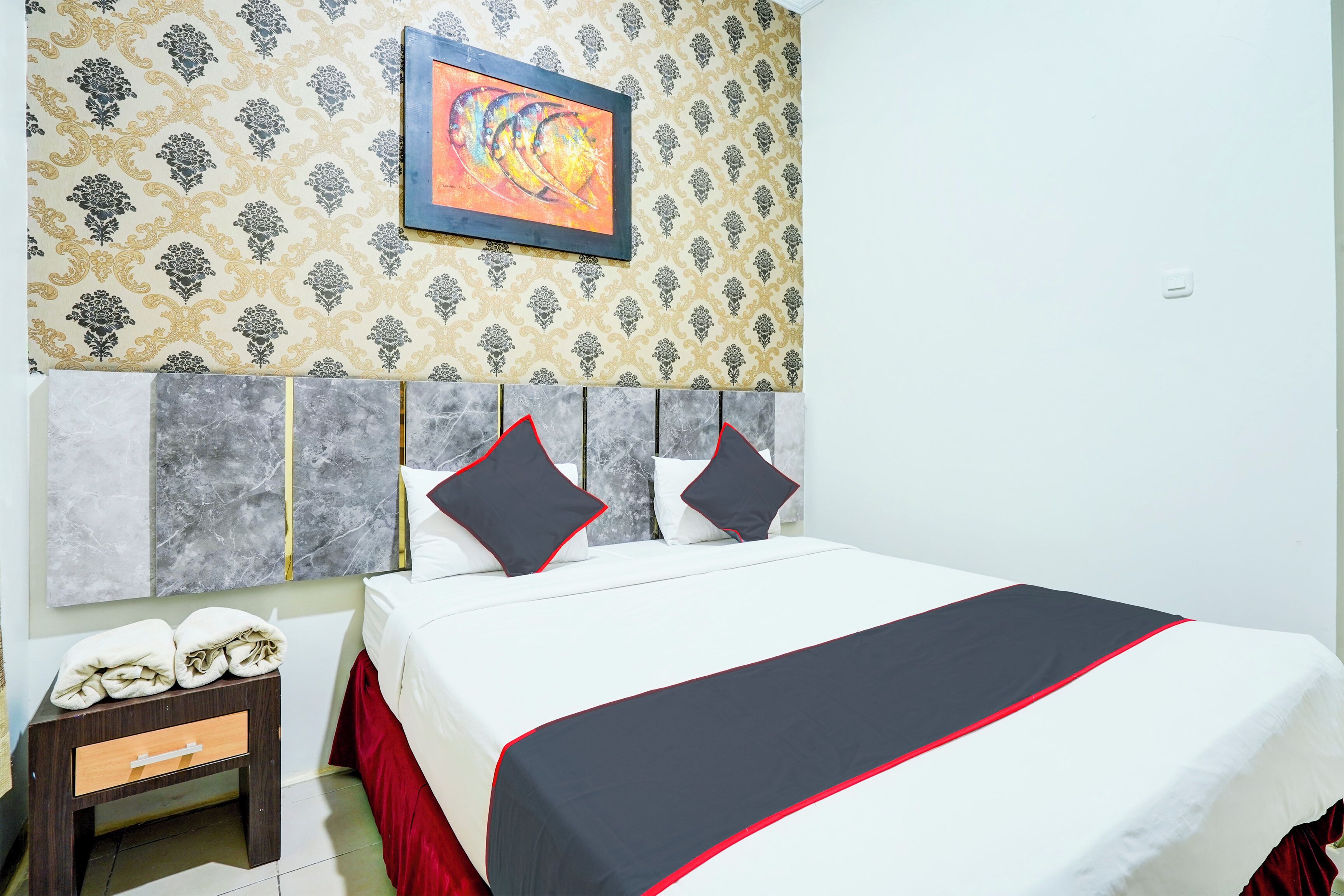 A neatly made bed with a white and black bedspread, two pillows, and a red skirted bed base. A small wooden nightstand with a drawer and a framed picture hangs on the wall above the bed.