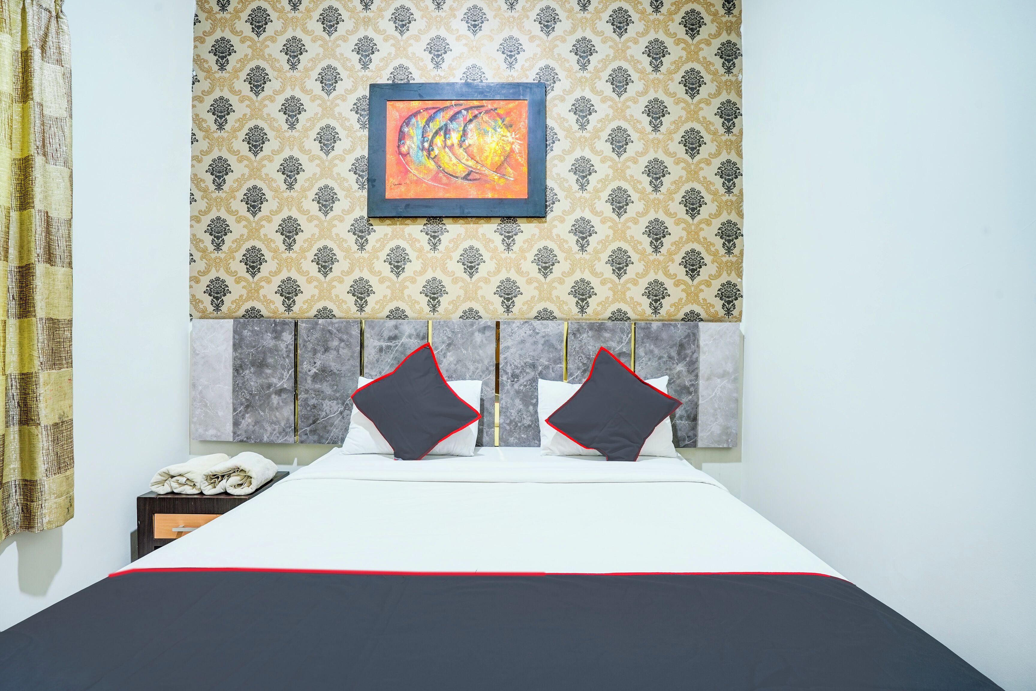 A hotel room with a bed, two pillows, a nightstand, a framed artwork, and patterned wallpaper.