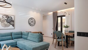 A modern living room with a sectional sofa, a dining table with chairs, a wall clock, and a ceiling light fixture.