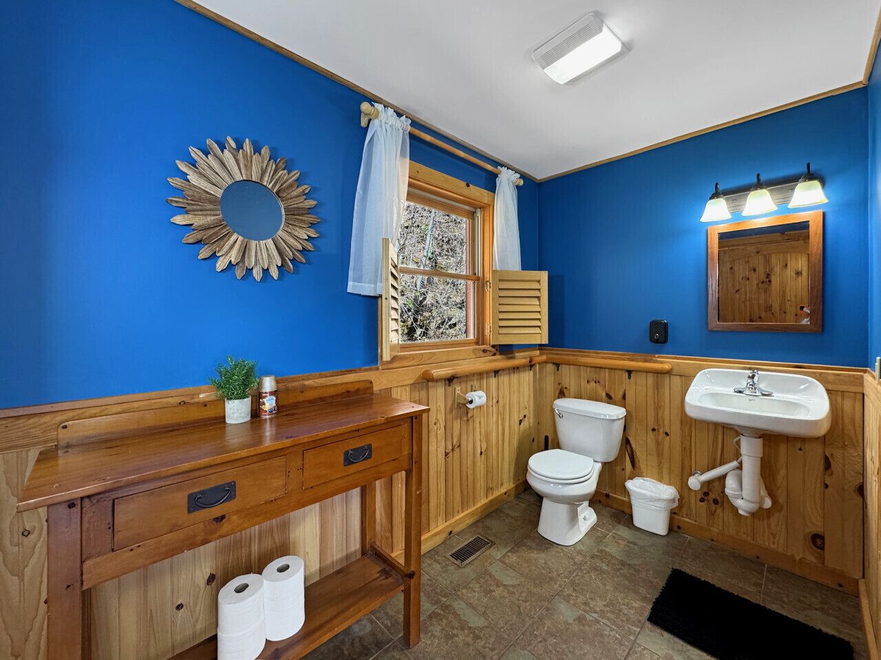 Une salle de bain avec des murs bleus, un lambris en bois, un meuble-lavabo en bois avec un miroir, des toilettes, un lavabo et une fenêtre en verre dépoli.
