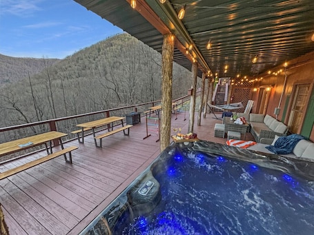 A hot tub on a wooden deck with benches and a sofa, overlooking a forested hillside.
