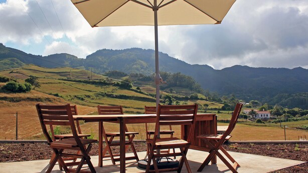 A table and chairs under a large umbrella with a view of rolling hills and a distant house.