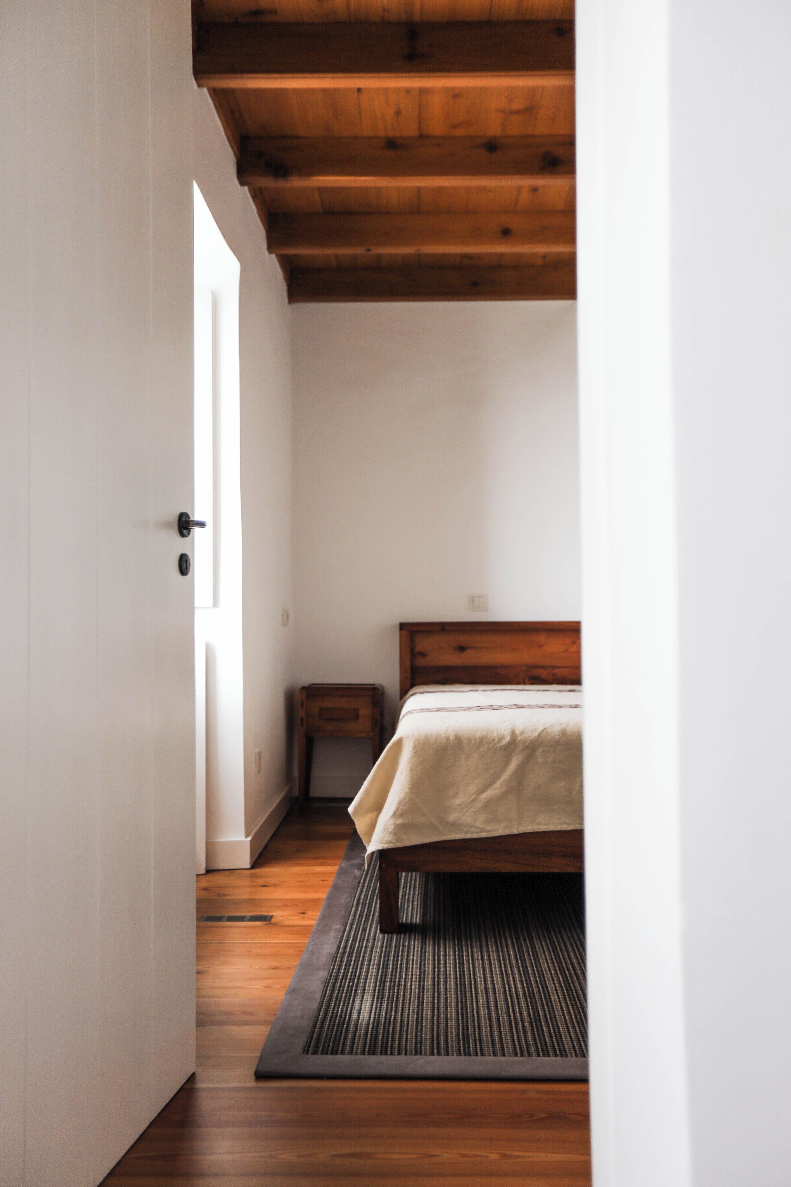 Une chambre avec un plafond en bois, un lit recouvert d’une couvre-lit beige, une petite table de chevet et un tapis foncé.