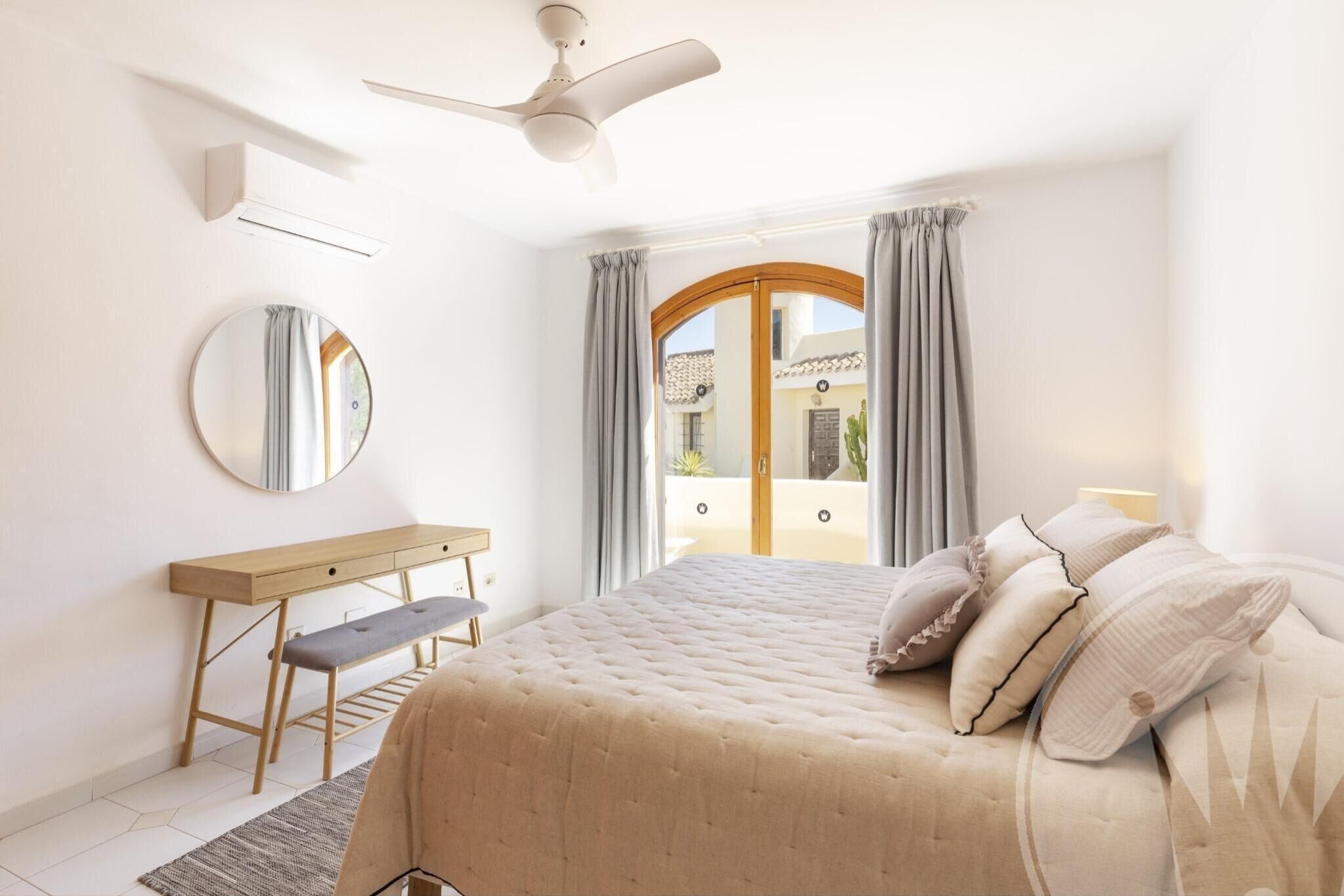 A bedroom with a bed, a desk, a chair, a round mirror, and a ceiling fan.