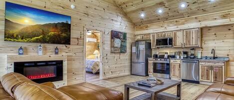 A cozy cabin interior with a fireplace, leather sofas, a kitchen, and a large TV.