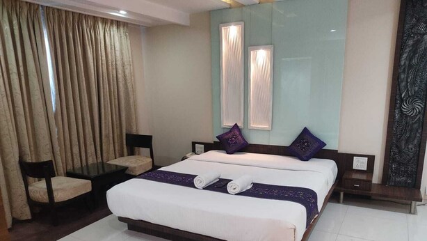 A hotel room with a large bed, a small table, a chair, and a window with curtains. - Sai Kan Hotel (Shirdi)