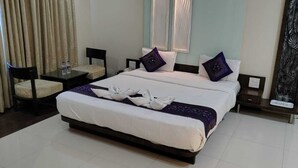 A hotel room with a neatly made bed, a chair, a small table with a lamp, and a decorative wall piece. - Sai Kan Hotel (Shirdi)