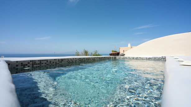 Una piscina azul cristalina con borde de azulejos, rodeada de arena blanca y vista al océano.