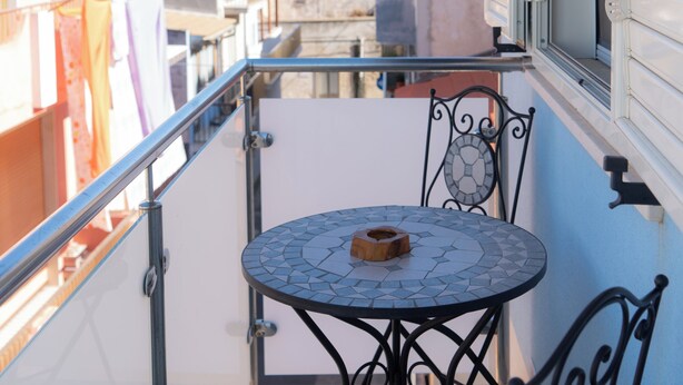 Un balcón con una mesa redonda y sillas, con vista a un paisaje urbano que incluye edificios y el mar.