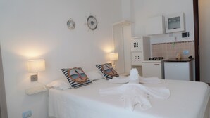 Habitación de hotel con una cama bien tendida, dos lámparas fijadas a la pared, una kitchenette y un reloj de pared.