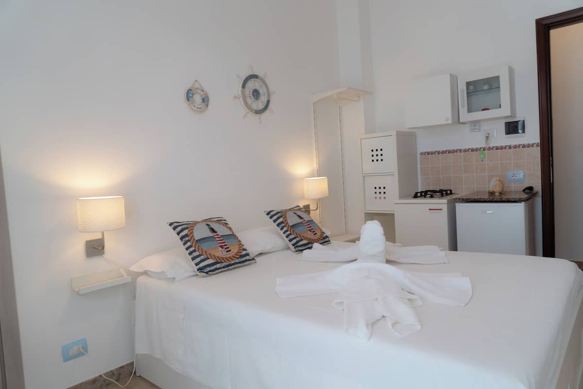 A hotel room with a neatly made bed, two wall-mounted lamps, a small kitchen area, and a wall clock.