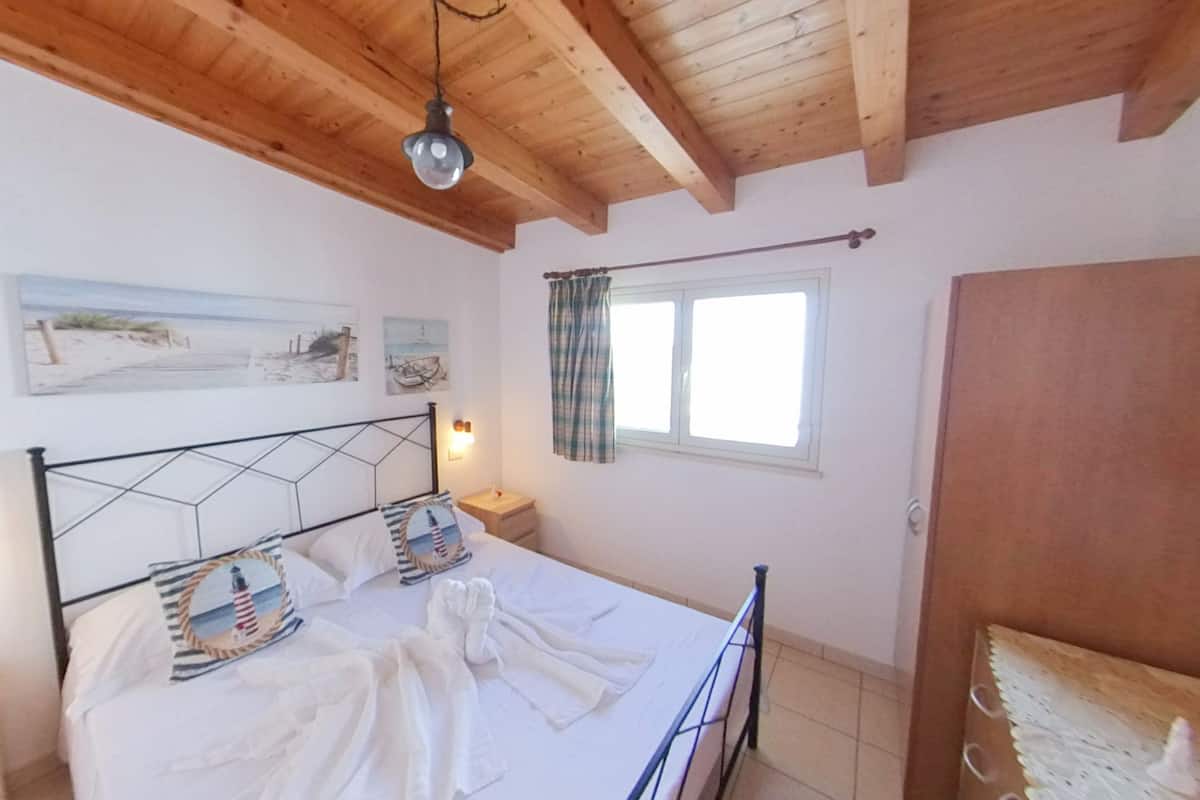 A bedroom with a bed, wooden ceiling beams, a window with curtains, and wall-mounted artwork.
