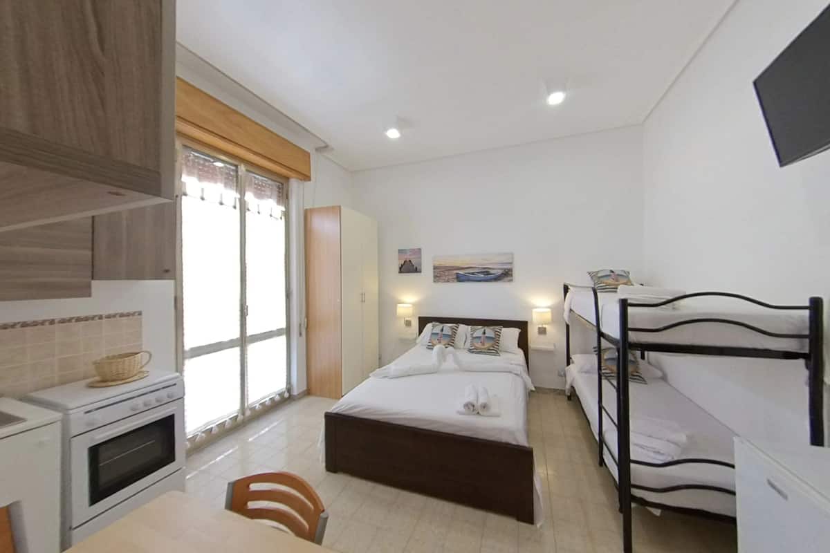 A compact hotel room with a kitchenette, a single bed with a headboard, a small table, and a window with curtains.