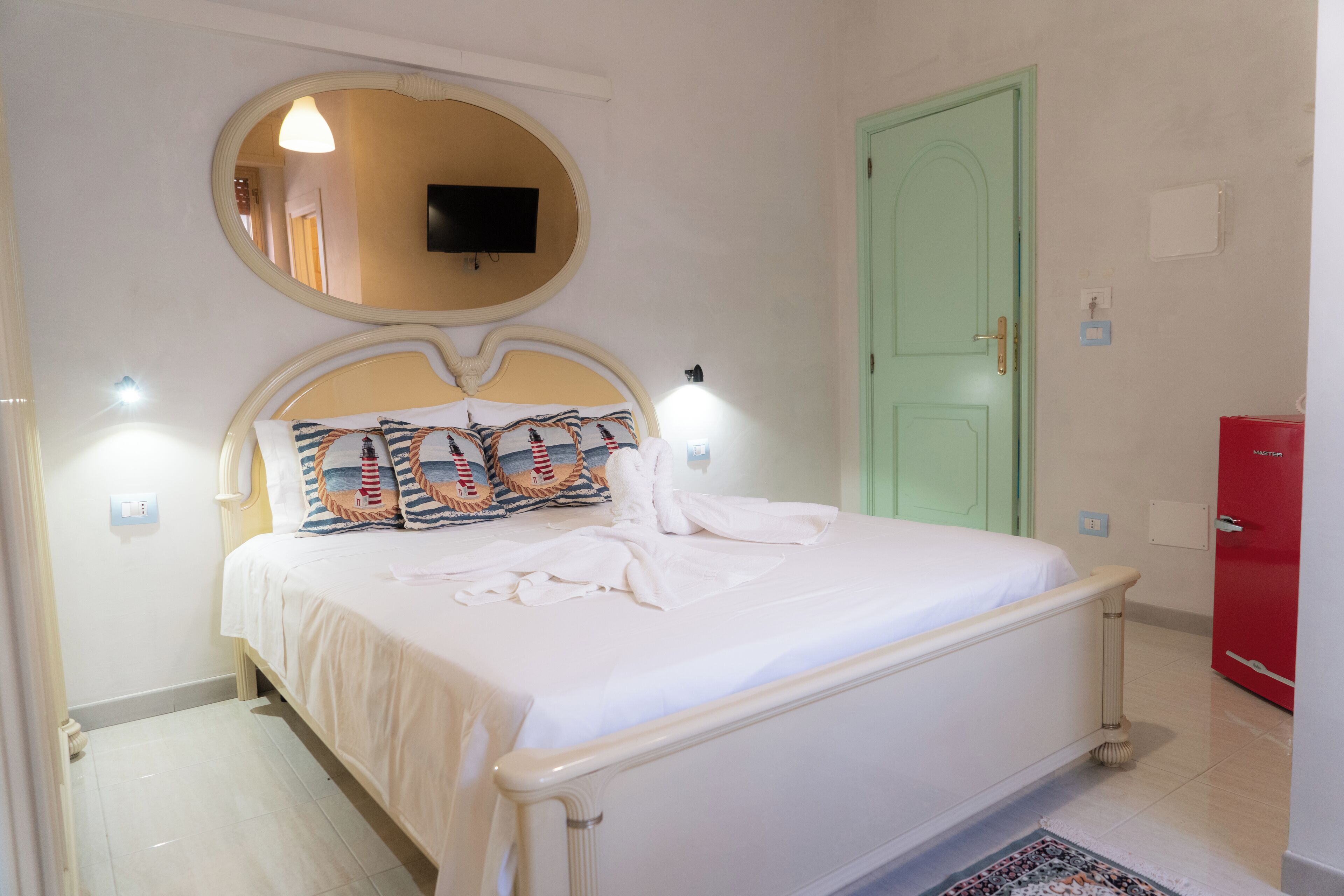 Ein ordentlich gemachtes Bett mit weißer Decke und Kissen, ein runder Spiegel darüber, eine grüne Tür und ein roter Schrank in einem Zimmer mit weißen Wänden und Fliesenboden.