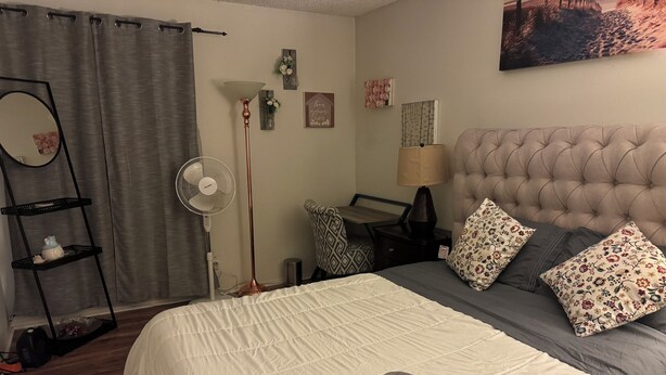 Una camera da letto ordinata con un letto, cuscini, un comodino, una lampada, un faretto da terra, uno specchio e una mensola a muro.