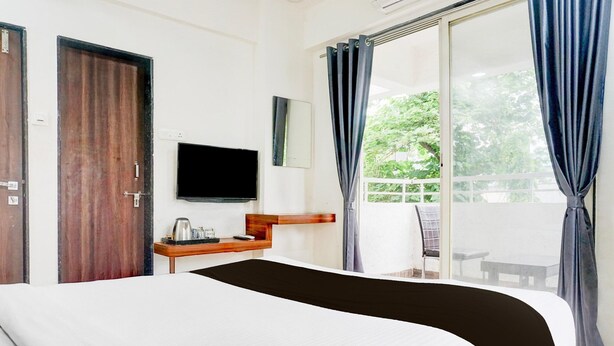 A hotel room with a bed, a television, a small table with a kettle, and a view of the outdoors through a large window.