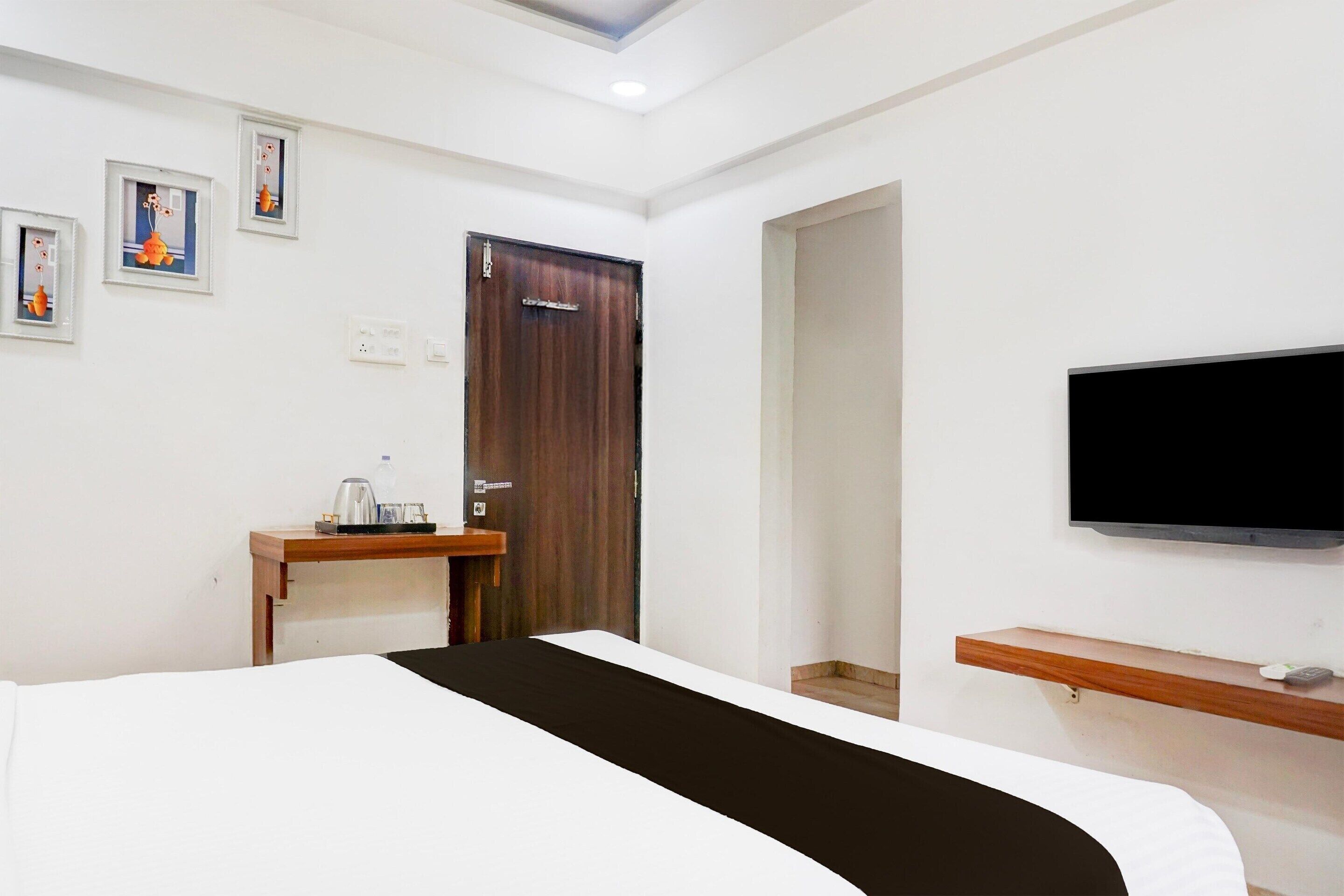A hotel room with a bed, a wooden nightstand with a kettle, a television mounted on the wall, and a door with a sign.