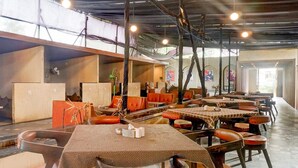 A spacious restaurant with high ceilings, exposed beams, and a long counter with orange cushioned chairs. Tables are covered with patterned tablecloths, and there are decorative plants and artwork on the walls.