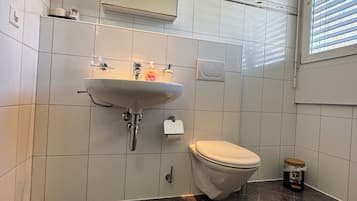 Un baño con lavamanos, inodoro y espejo. Tiene una ventana con persianas y un pequeño cubo de basura.