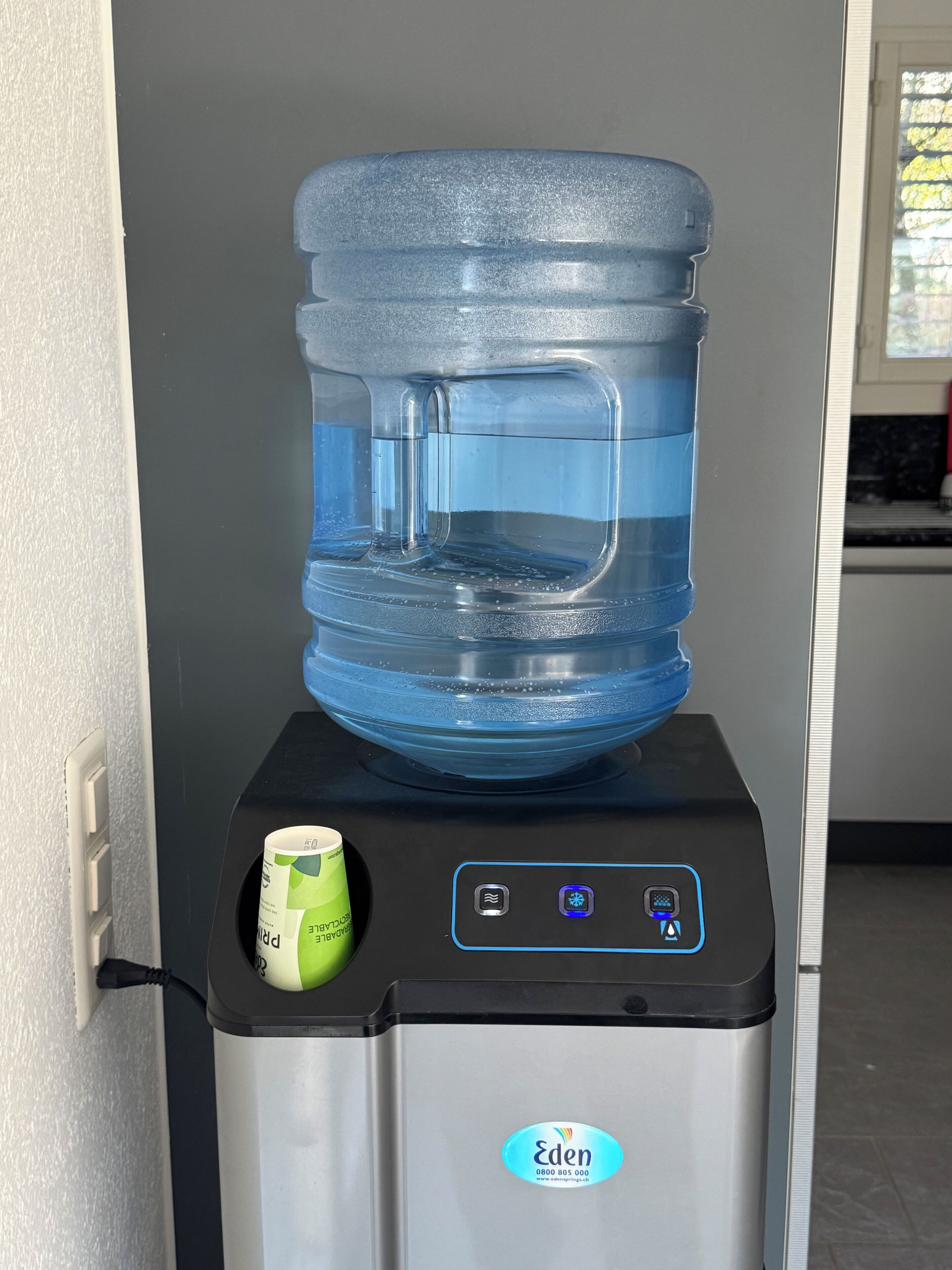 A water dispenser with a large water jug on top, a green cup, and a control panel with a brand logo.