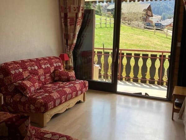 A room with a patterned sofa, a wooden floor, and a view of a rural landscape through a glass door.