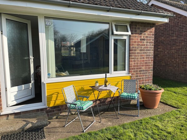 A sunlit room with a yellow wall, a sliding glass door, and a window. There are two chairs and a small table with a bottle and glass.