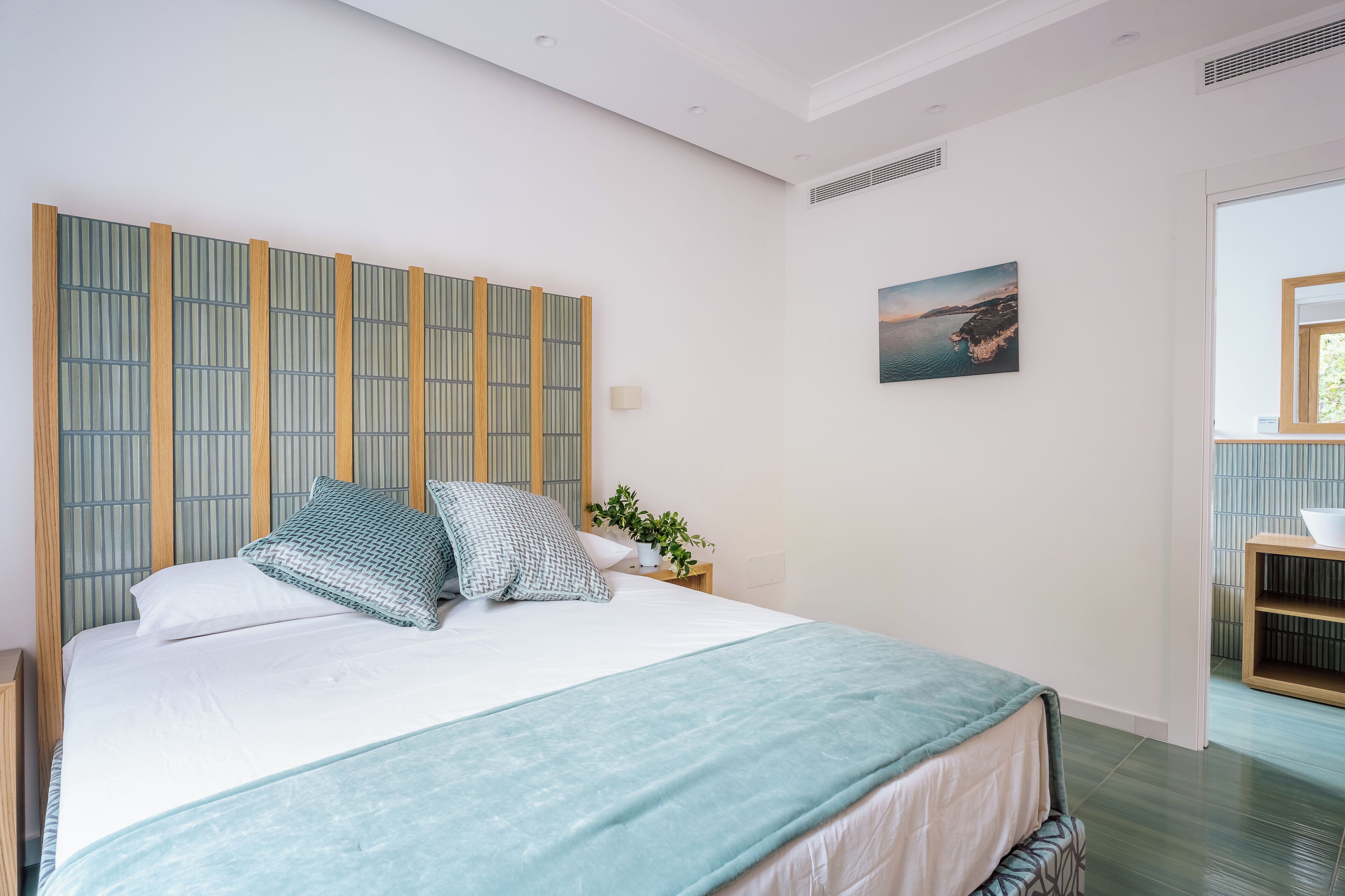 Un dormitorio ordenado con cama, almohadas, una mesita de noche con una planta y un cuadro colgado en la pared.