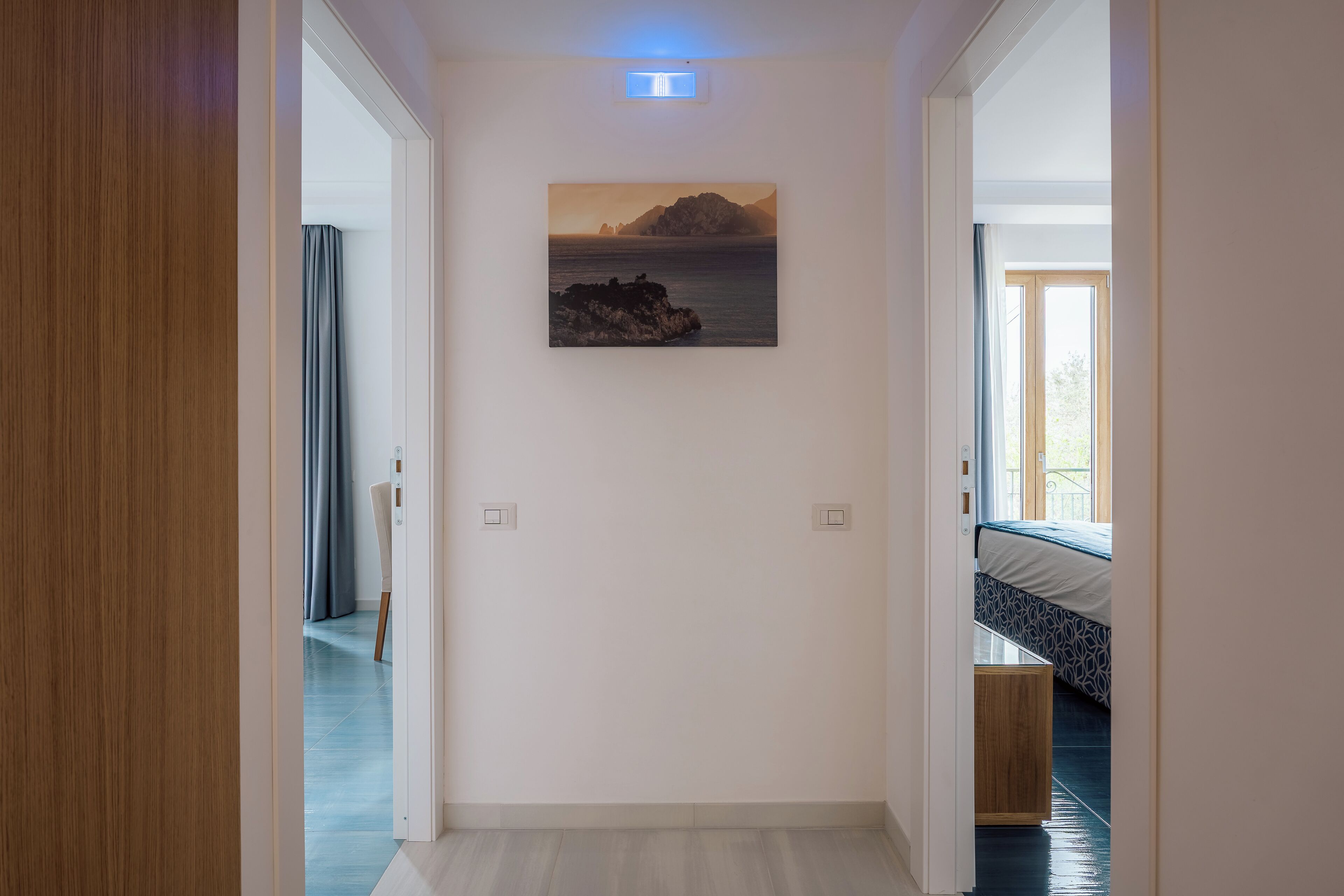 Un couloir orné d’un tableau représentant un paysage côtier, un lit recouvert d’une couvre-lit à motifs et une porte en bois.
