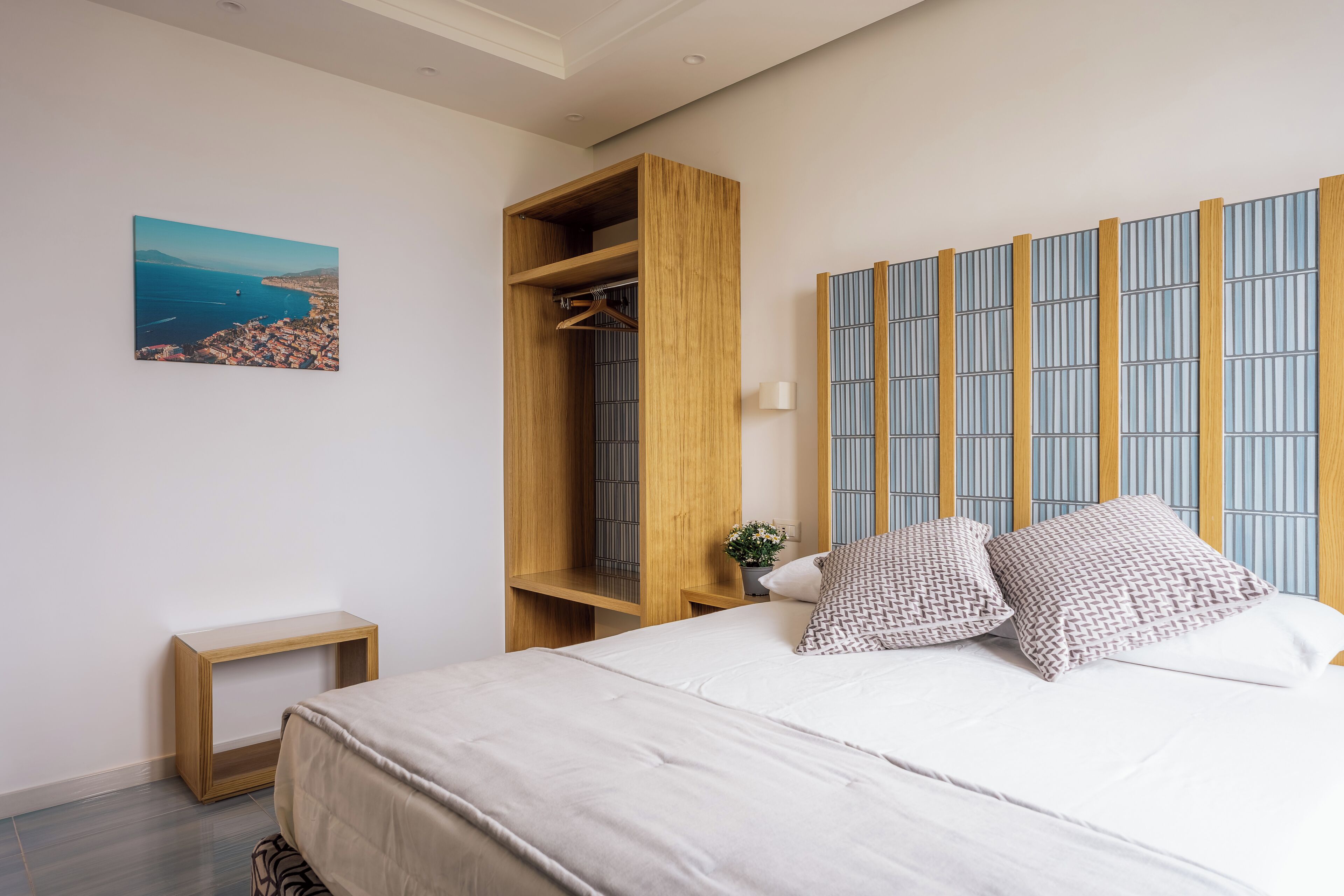 Quarto com cama, guarda-roupa de madeira, mesinha de cabeceira, luminária na parede e pintura com vista para a cidade.
