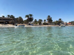 A beach with clear turquoise water, several boats, palm trees, and beachside buildings.