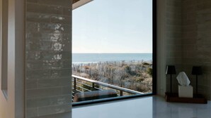 Habitació amb finestra gran que dona a una platja i oceà, amb pared de maó, barana i dues làmpades.