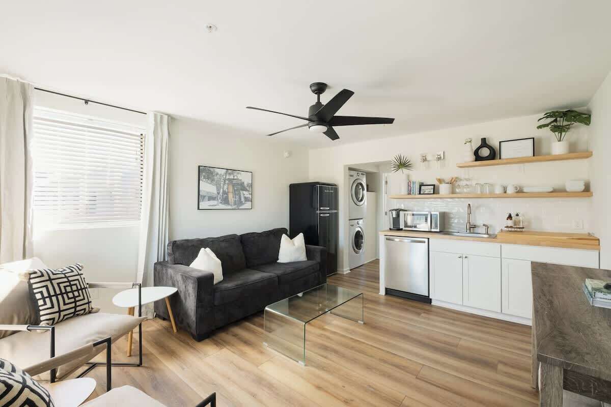 A modern living room with a black sofa, a glass coffee table, and a dining area with a wooden table. There is a kitchen with a microwave, oven, and washing machine. The room has wooden flooring and white walls.