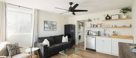 A modern living room with a black sofa, a glass coffee table, and a dining area with a wooden table. There is a kitchen with a microwave, oven, and washing machine. The room has wooden flooring and white walls.