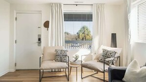 Una sala de estar moderna con dos sillones color beige, una mesita redonda y un sofá oscuro. Un ventanal con persianas y cortinas deja entrar luz natural.