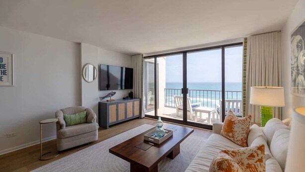 A living room with a sofa, coffee table, and a TV on a wall. There is a balcony with a view of the ocean.