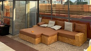 Un espace de détente extérieur moderne, doté d’une structure en verre, d’un banc rembourré et offrant une vue sur le quartier environnant.