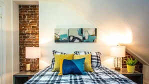 Une chambre avec une couvre-lit à carreaux, un mur en briques, des œuvres d’art abstraites et deux lampes de chevet.
