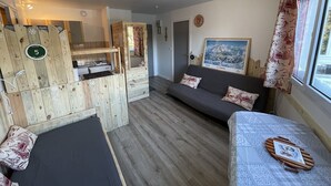 A compact room with a bed, a sofa, a wooden partition with a small kitchenette, and a window with curtains. - Cozy T2 in the heart of Le Lioran (Le Lioran)