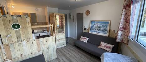 Een compacte kamer met een bed, een slaapbank, een houten scheidingswand met een kleine kitchenette en een raam met gordijnen.