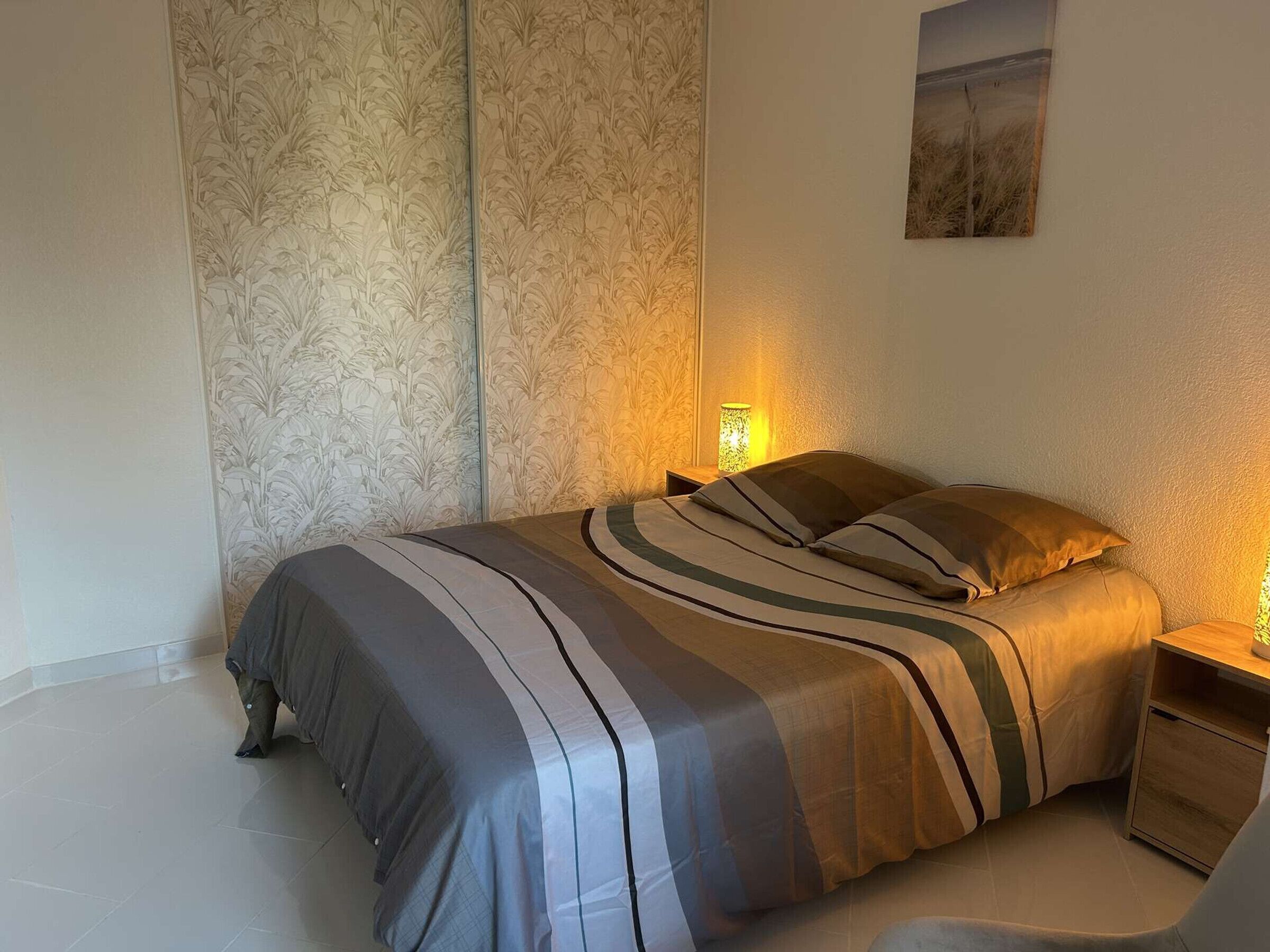 Un lit bien fait, avec une literie à rayures, une table de chevet avec une lampe et un mur recouvert d’un papier peint à motifs floraux.