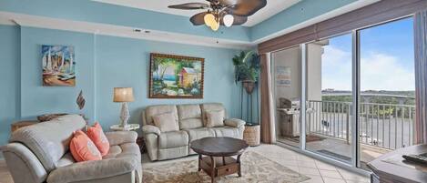 A living room with a sofa, armchair, coffee table, ceiling fan, artwork, and a balcony with a view.