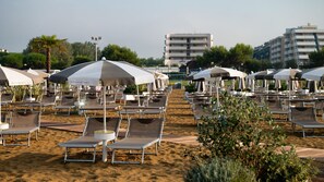 A beach with rows of sun loungers and umbrellas, a sandy beach, and buildings in the background.