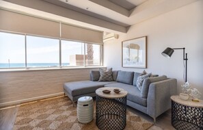 Ein modernes Wohnzimmer mit einer grauen Eckcouch, einem runden Couchtisch und einem Beistelltisch. Ein großes Fenster bietet Meerblick und führt auf einen Balkon.