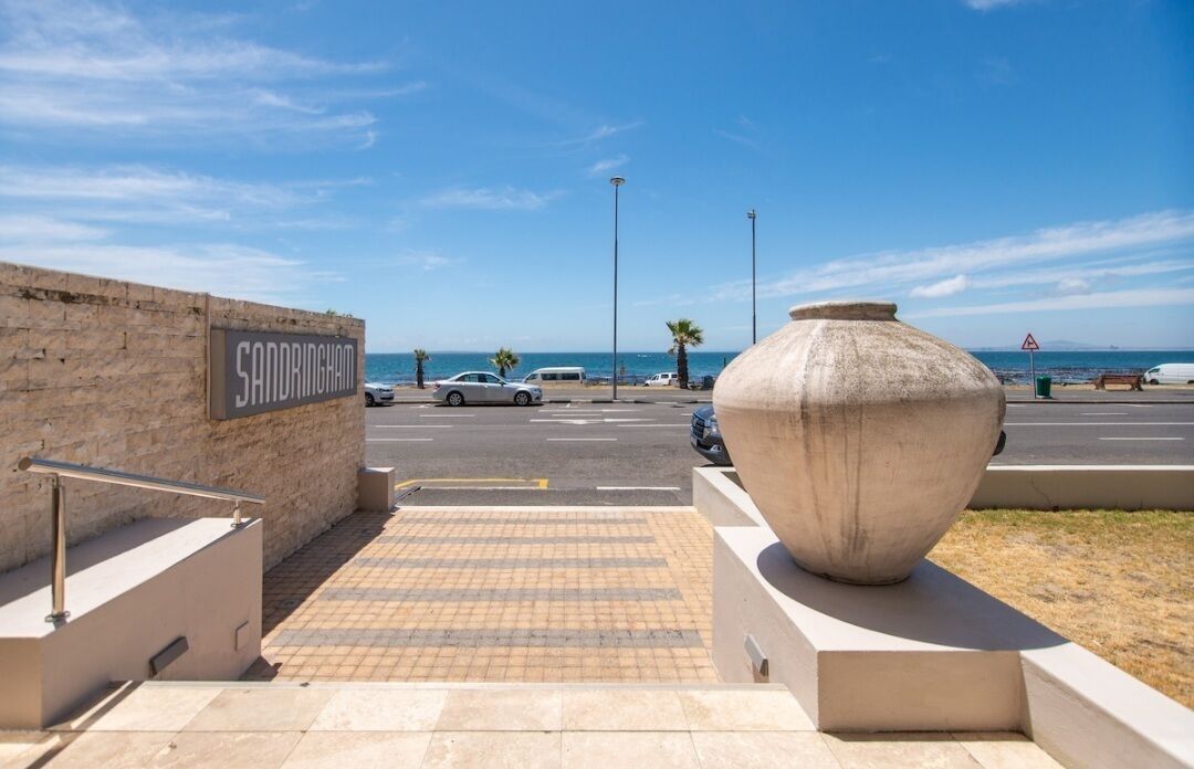 A parking area with a large concrete planter, a sign reading "SANDRINGHAM", and a clear view of the beach and ocean in the background.