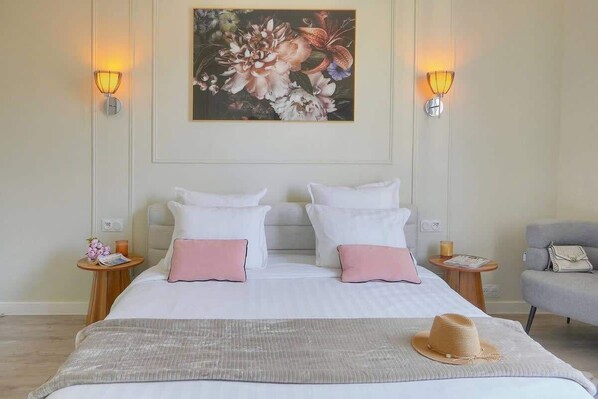 A double bed with white and pink pillows, a beige throw blanket, and a straw hat on the bed. There are two bedside tables with lamps, a gray armchair, and a painting on the wall. - EMILY 2BR 2baths with AC near the Palais (Cannes)